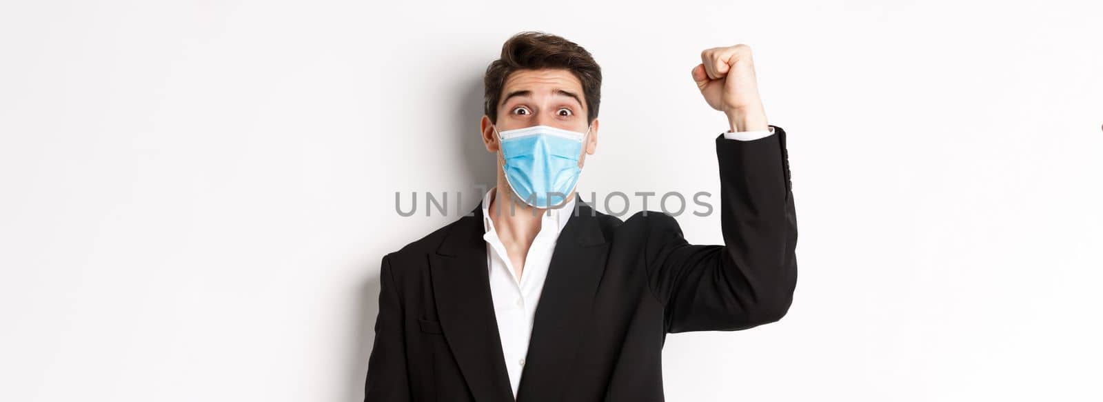 Concept of covid-19, business and social distancing. Close-up of excited businessman in medical mask and suit, rejoicing, achieving goal and triumphing, white background by Benzoix
