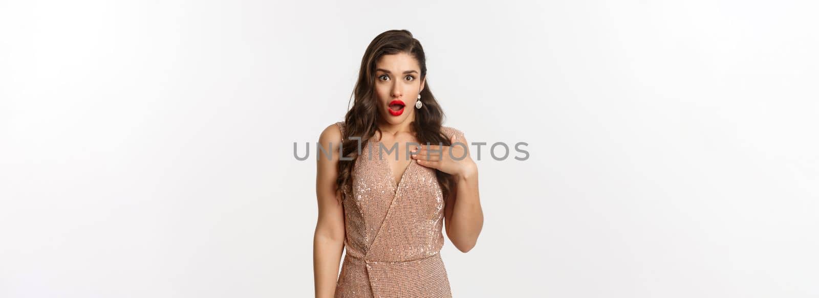 Winter holidays and party concept. Image of attractive woman with red lips, wearing glamour dress and pointing at herself with shock, looking surprised, standing over white background.