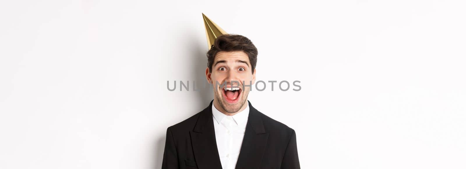 Close-up of excited handsome guy in trendy suit and party hat, looking amazed, celebrating winter holidays, standing against white background by Benzoix