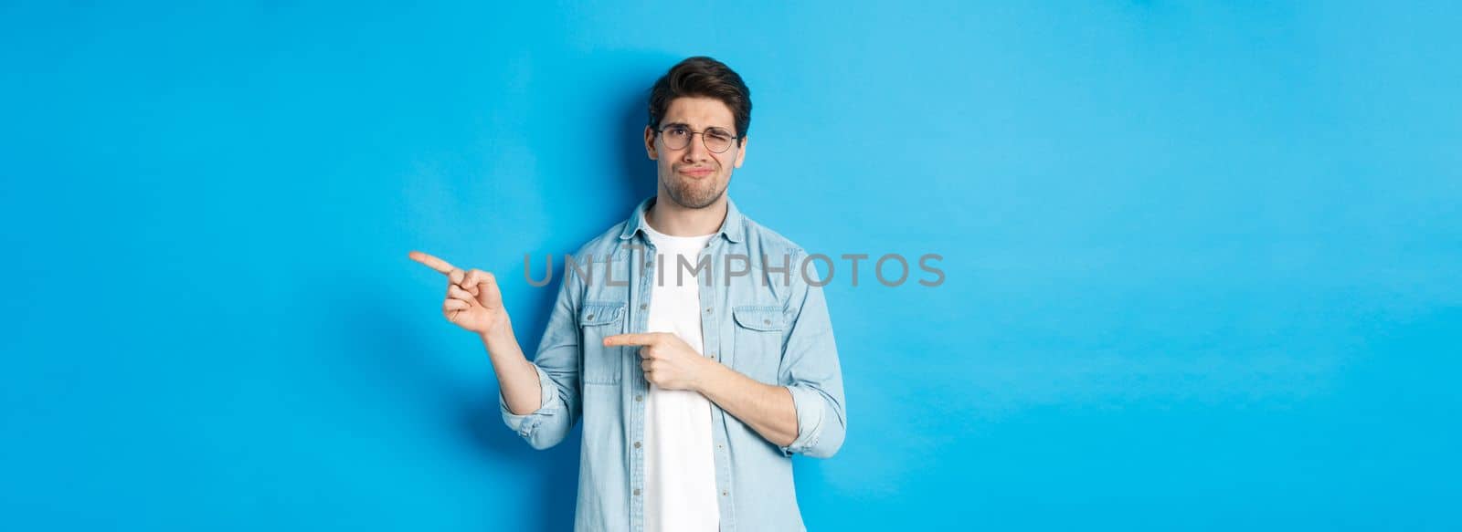 Disappointed young man in glasses pointing fingers right at copy space, showing promo banner and smirking dissatisfied, judging bad product, standing over blue background by Benzoix