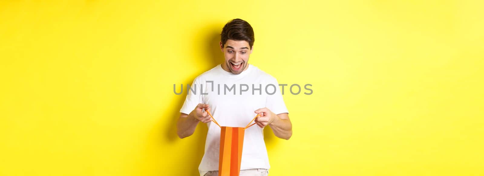 Excited guy open bag with gift, looking inside with amazement and happy face, standing against yellow background.
