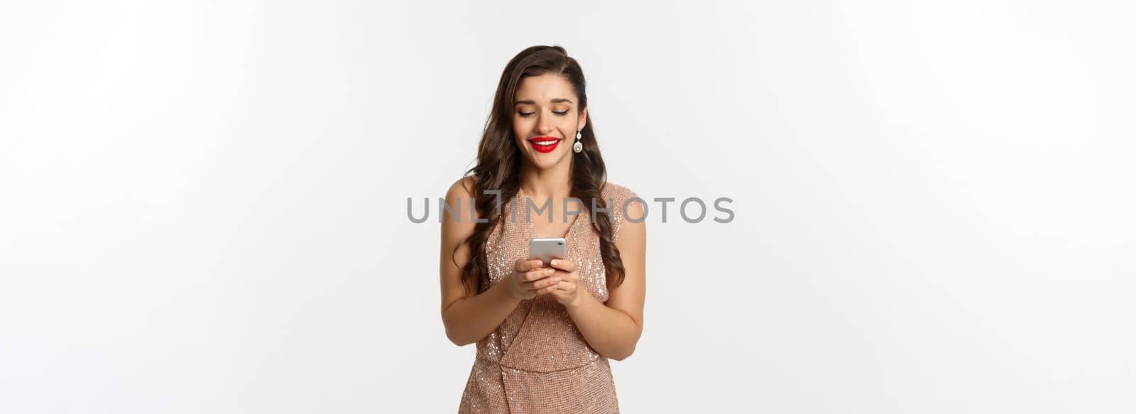 Christmas party and celebration concept. Beautiful woman in glamour dress reading text message on phone and smiling, using smartphone, standing over white background.