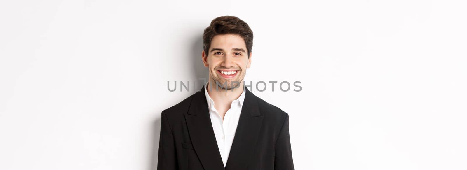 Close-up of handsome young businessman in trendy suit smiling, standing against white background.