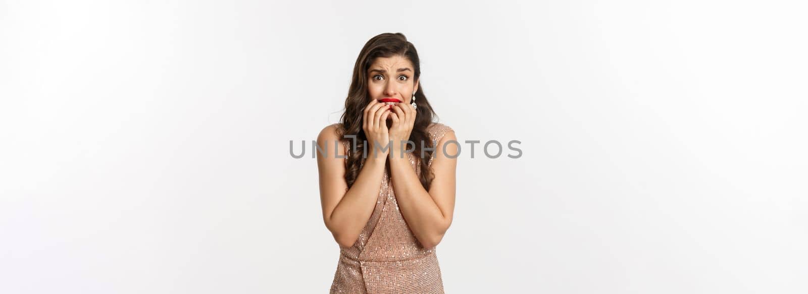 Celebration and party concept. Scared woman biting fingernails and anxiously staring at camera, standing worried in glamour dress, white background by Benzoix