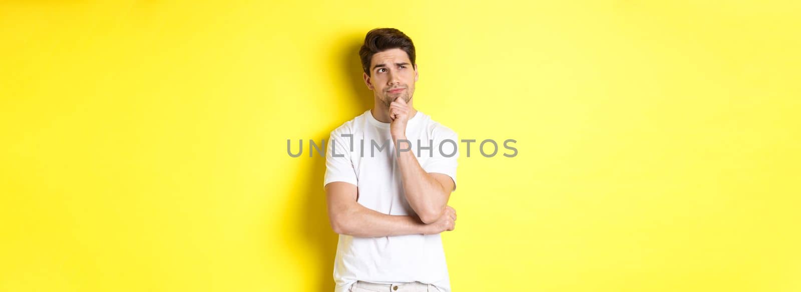 Portrait of young male model thinking, looking at upper left corner and making choice, standing near copy space, yellow background by Benzoix