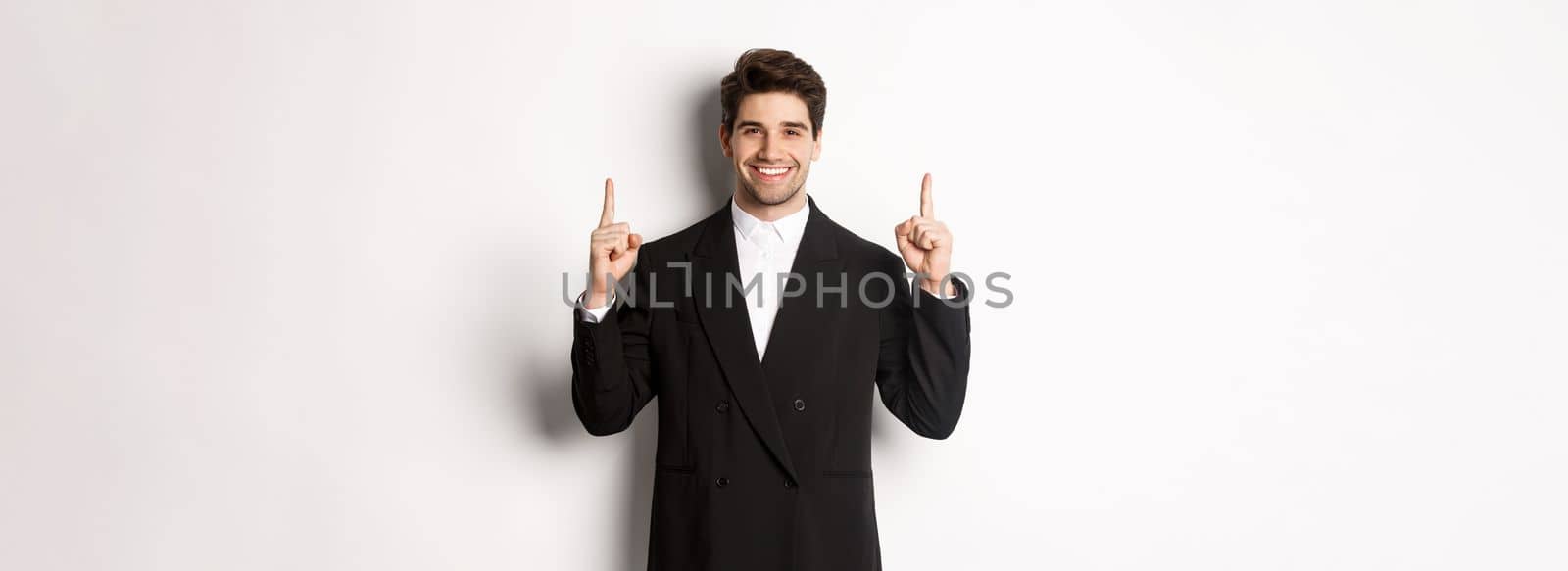 Portrait of attractive caucasian man in stylish black suit, pointing fingers up and smiling, showing christmas advertisement, standing over white background by Benzoix