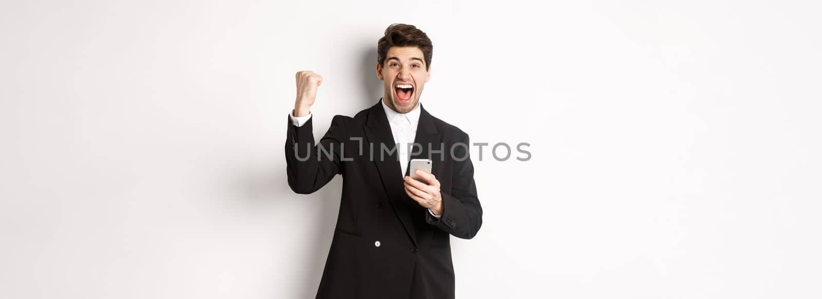 Portrait of happy handsome man in suit, rejoicing, achieve goal on mobile app, raising fist up and shouting yes, holding smartphone, standing against white background by Benzoix