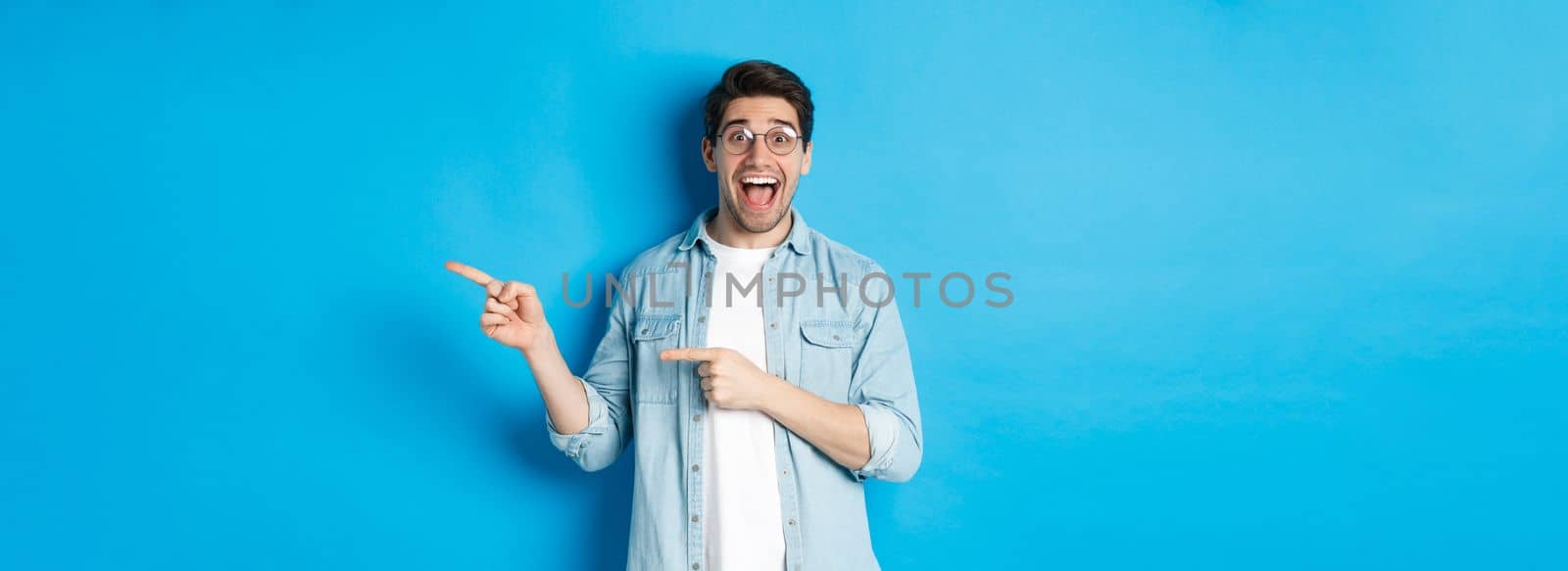 Surprised man in glasses pointing right at copy space, showing promo offer on blue background, standing over blue background.