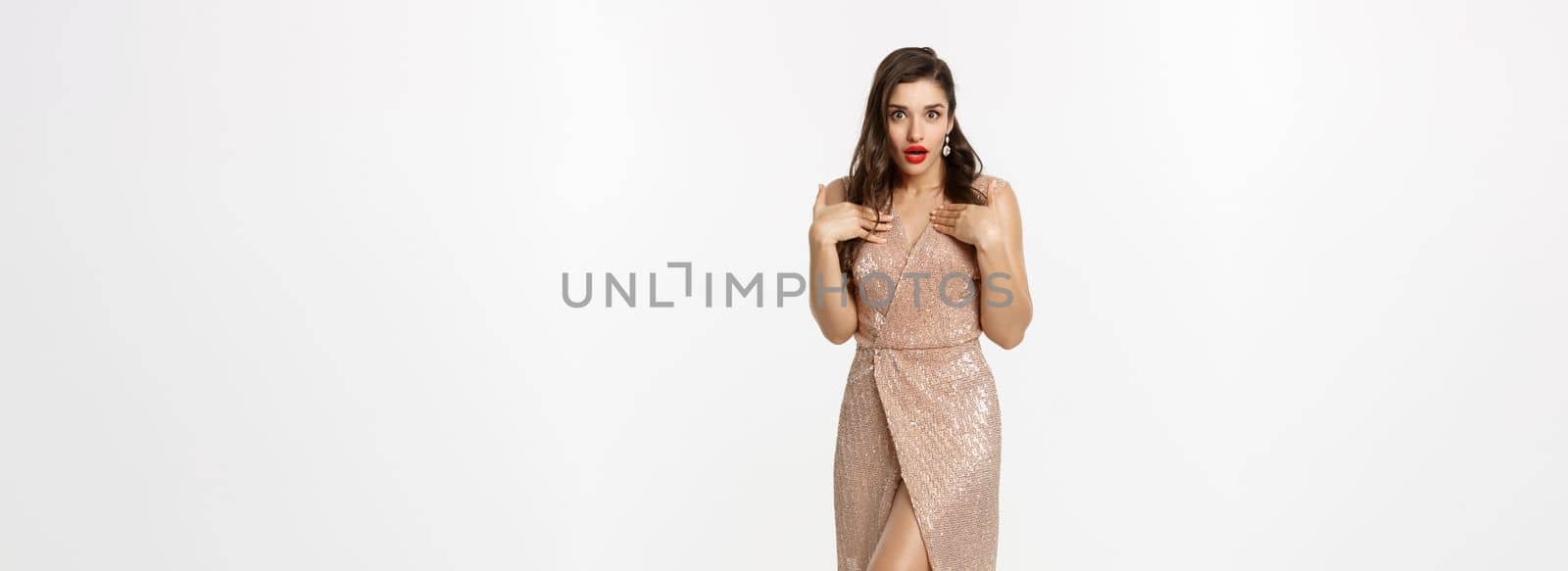 Christmas party and celebration concept. Full length of attractive female model in elegant dress, looking surprised and pointing at herself, standing over white background.