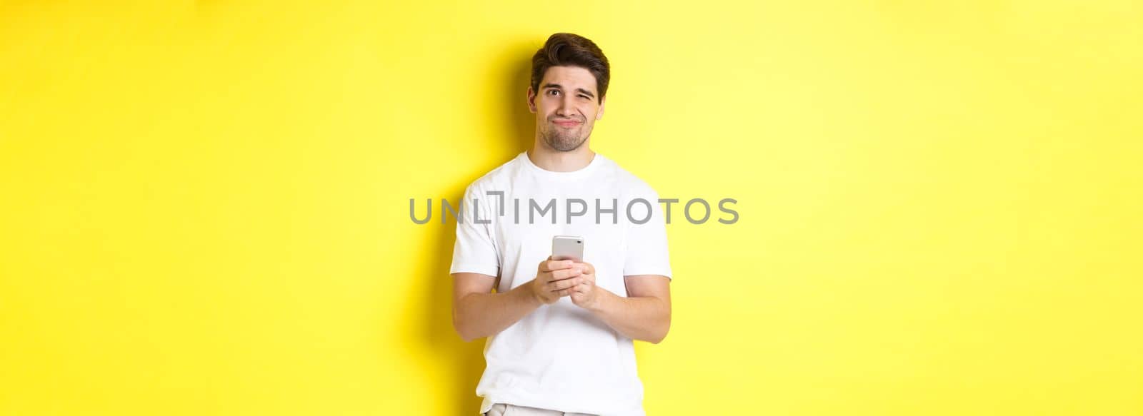Displeased and reluctant man grimacing, being unamused by message on smartphone, standing over yellow background.