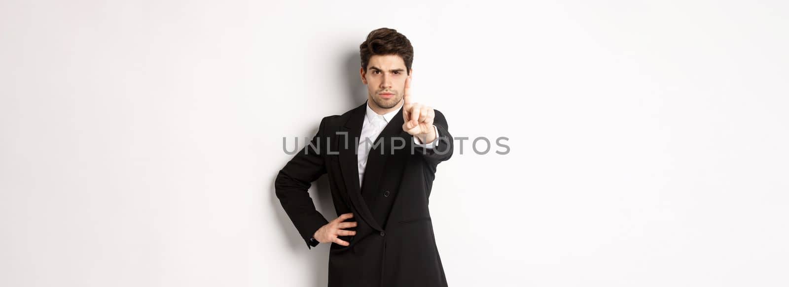 Portrait of serious handsome man in business suit, showing one finger to prohibit or decline something, telling to stop, disagree with you, standing over white background by Benzoix