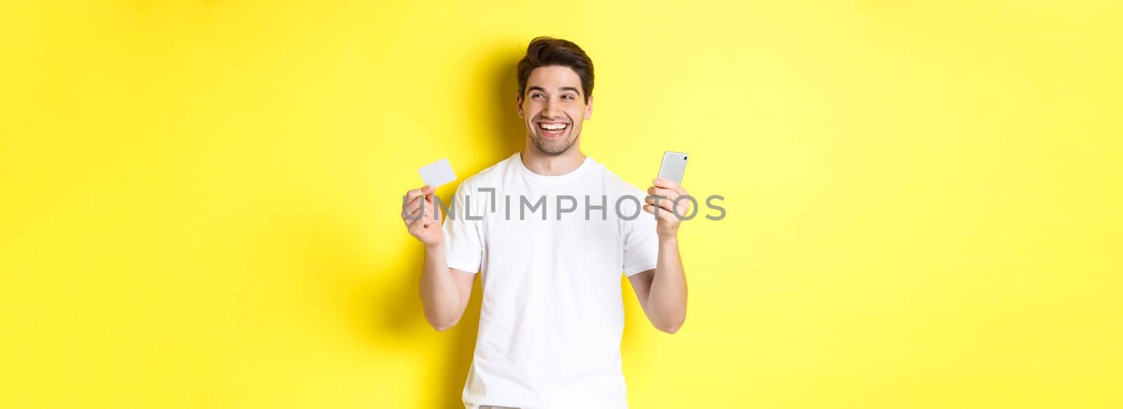 Happy man thinking about shopping, holding credit card and smartphone, smiling pleased, standing over yellow background.