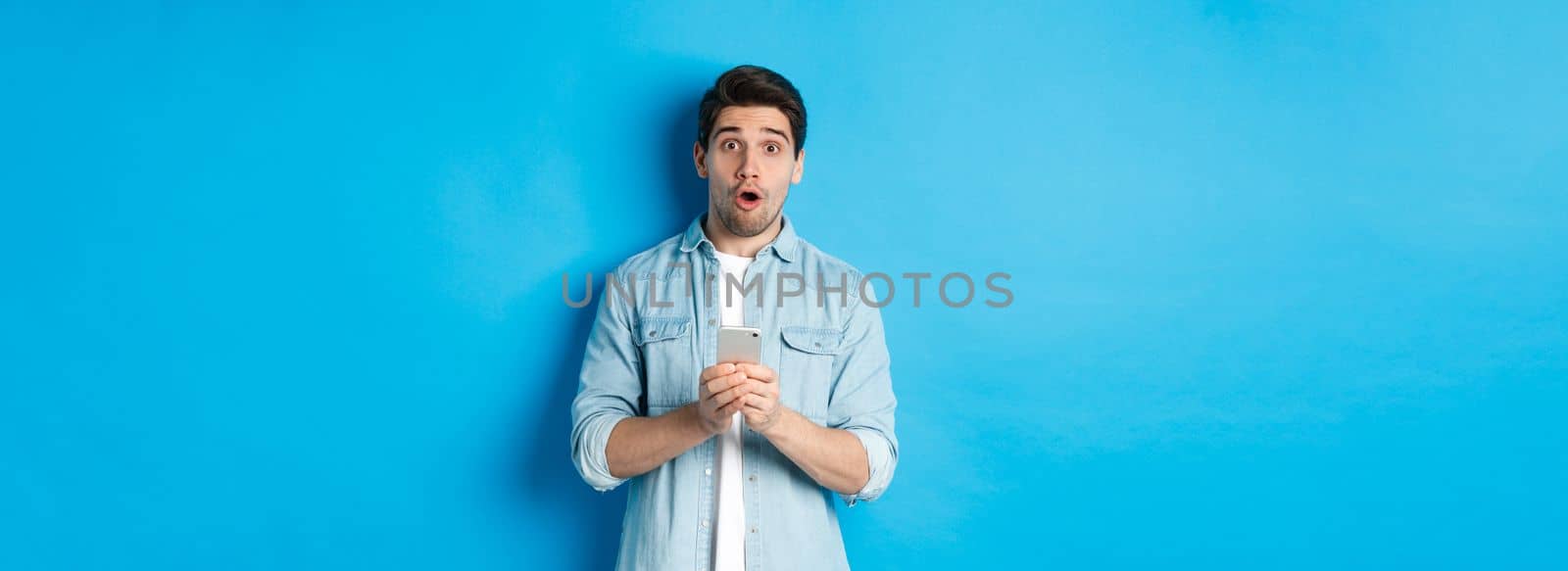 Surprised man in casual clothes looking astounded, holding smartphone, standing against blue background.