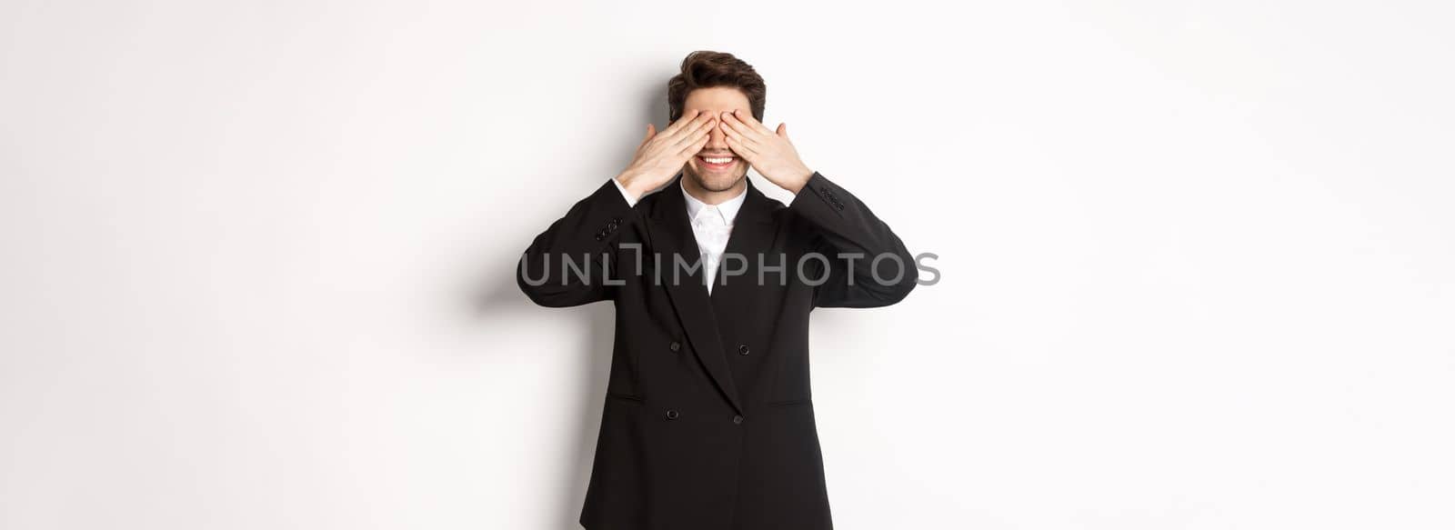 Image of handsome stylish man in black suit, waiting for christmas surprise, covering eyes with hands and smiling, anticipating presents, standing over white background.