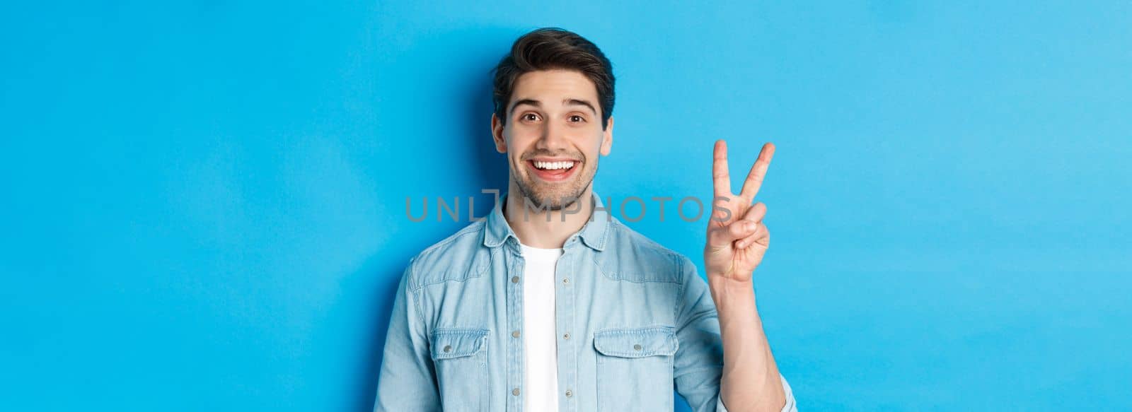 Close-up of handsome man smiling, showing fingers number two, standing over blue background.
