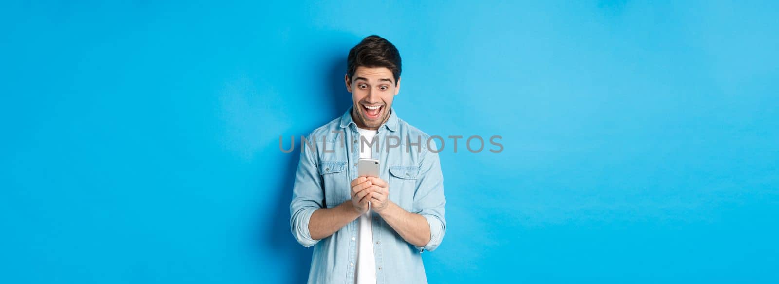 Image of excited man smiling while looking at mobile phone, shopping online on smartphone, standing against blue background by Benzoix