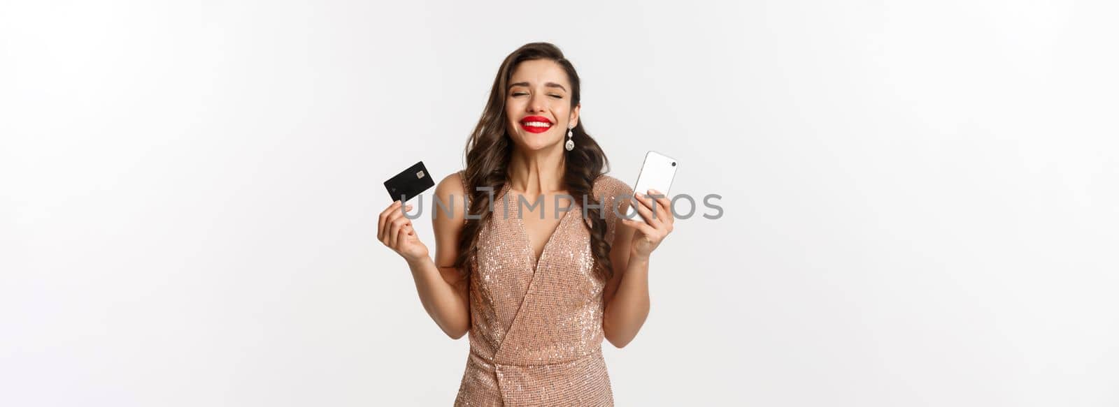 Online shopping and holidays concept. Satisfied and happy woman in elegant dress smiling, using credit card and mobile phone, standing over white background by Benzoix