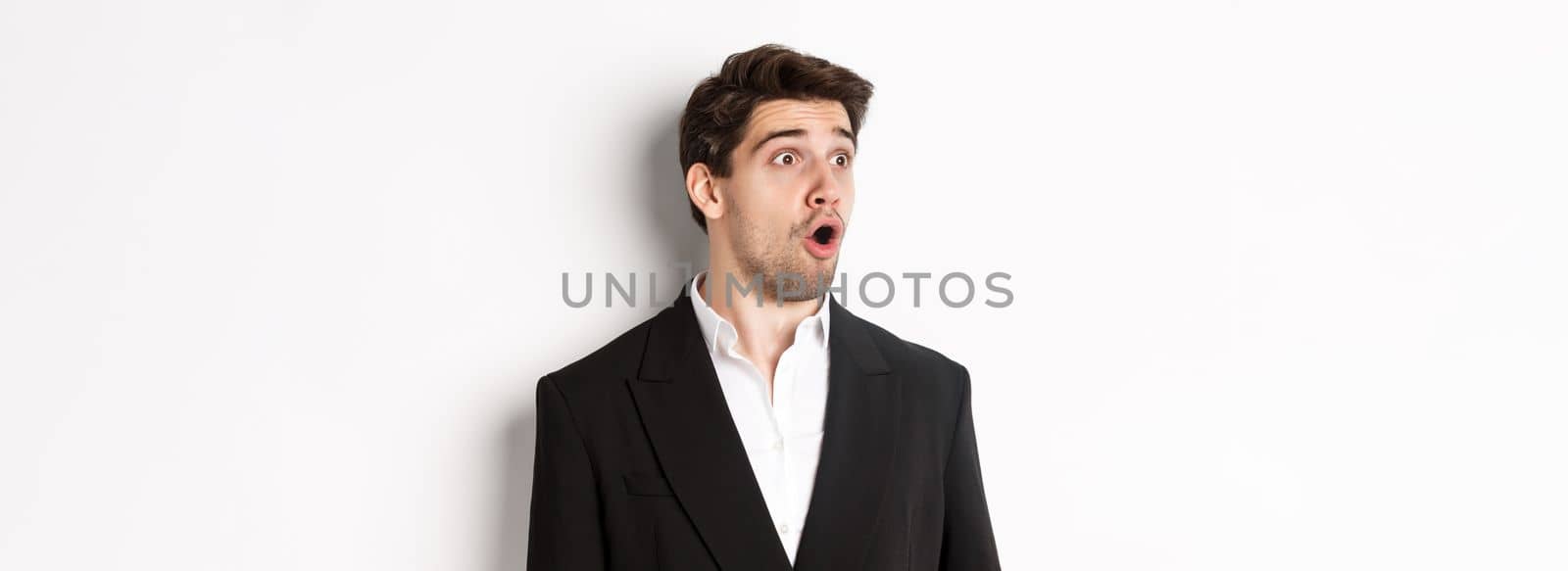 Close-up of impressed guy in trendy suit, open mouth and looking left wondered, standing against white background.