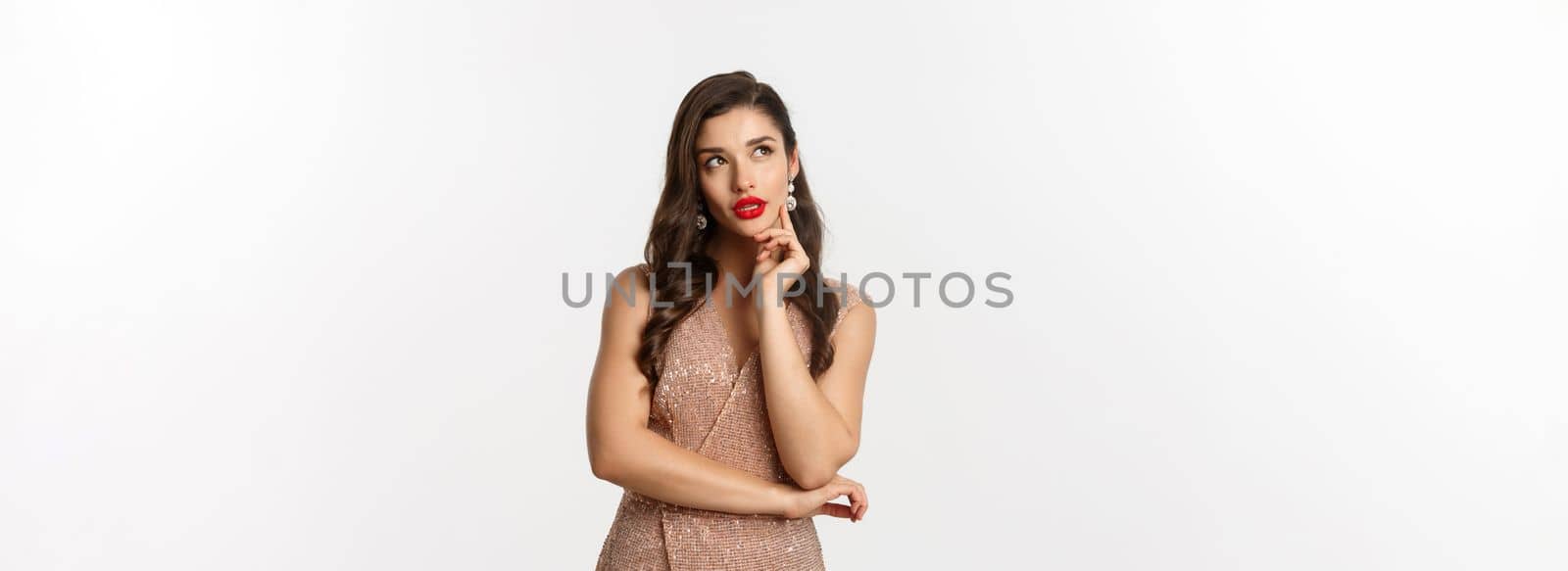 Christmas, holidays and celebration concept. Young woman looking thoughtful at upper left corner, wearing evening dress for party, thinking about New Year eve, white background.