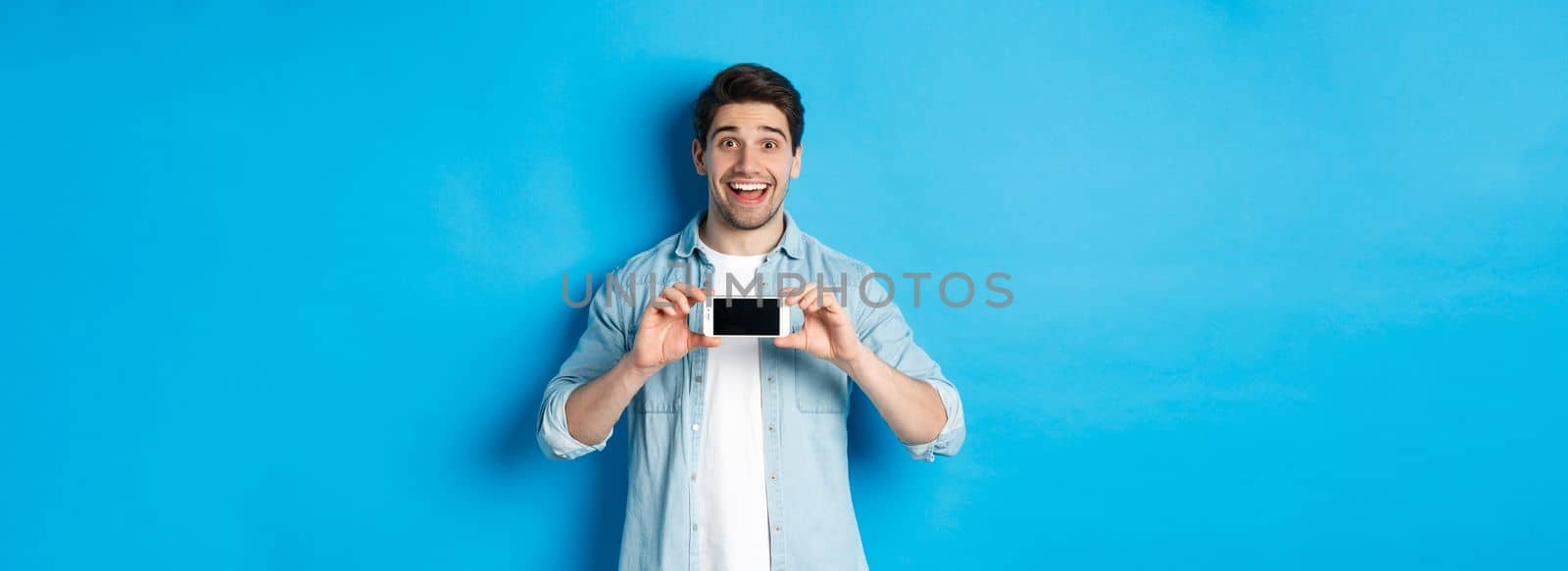 Amazed smiling man showing smartphone screen, internet promo offer, standing against blue background by Benzoix