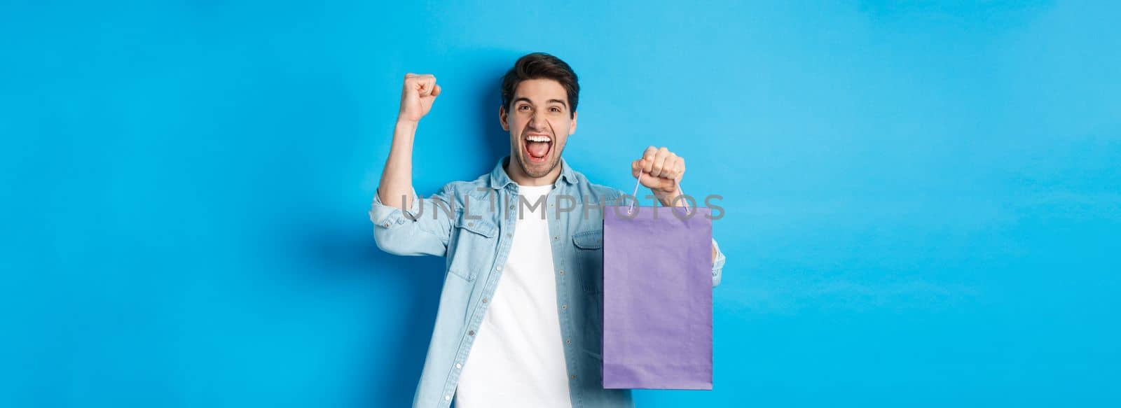 Concept of shopping, holidays and lifestyle. Cheerful young man celebrating, holding paper bag and making fist pump like winner, standing over blue background.