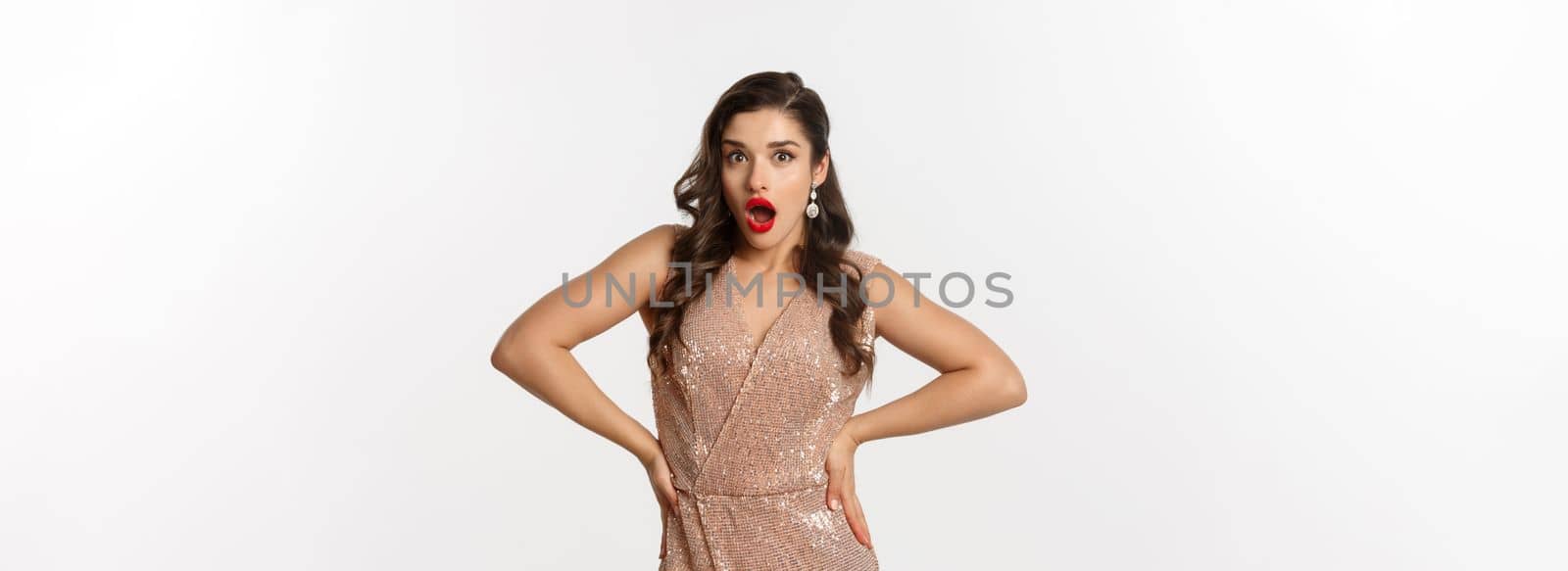 Image of attractive party woman in elegant dress, red lips, looking amazed and surprised, celebrating new year, standing over white background by Benzoix
