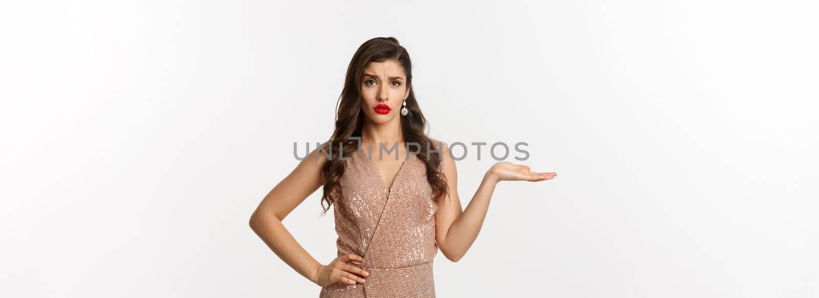 Unbothered young woman in elegant dress dont understand whats wrong, raising hand and looking careless, standing over white background.