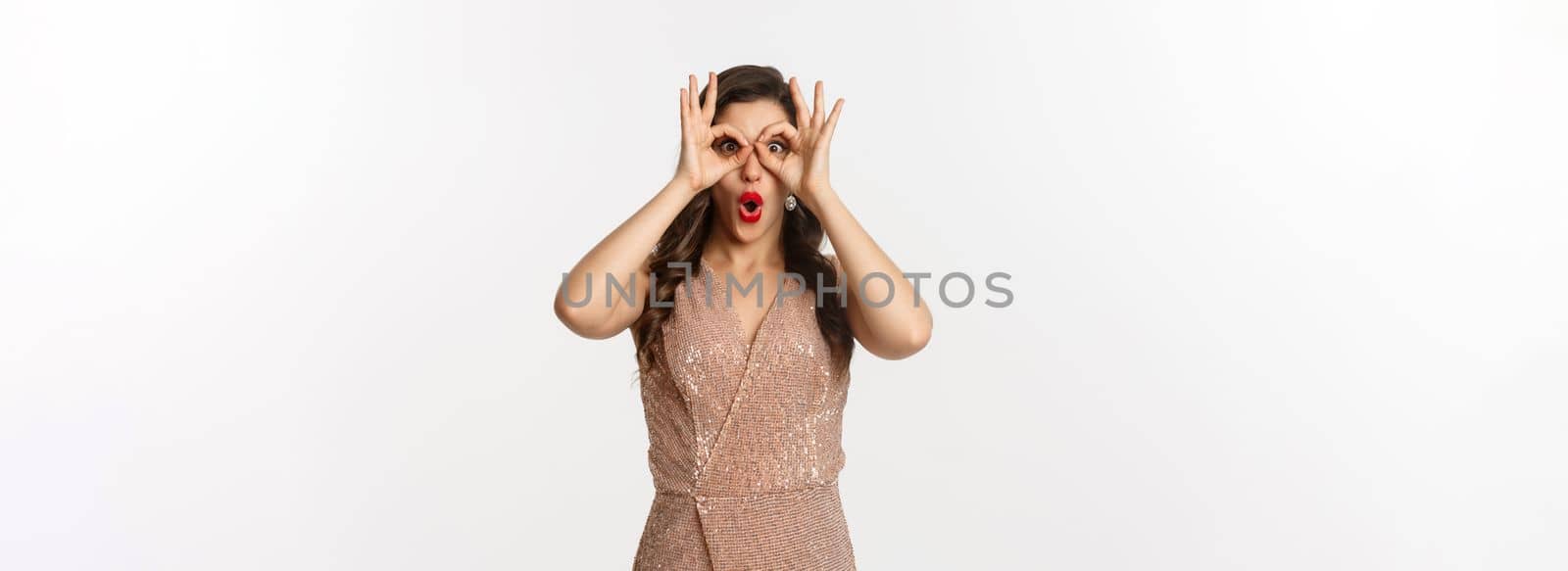 Christmas party and celebration concept. Woman in glamour dress and red lips looking at something amazing, staring through hand binoculars, standing over white background by Benzoix