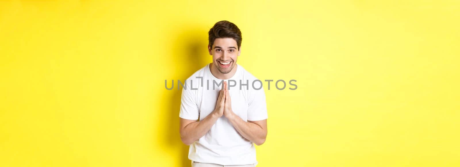 Handsome guy saying thank you, bowing and holding hands in namaste gesture, express gratitude, standing over yellow background.