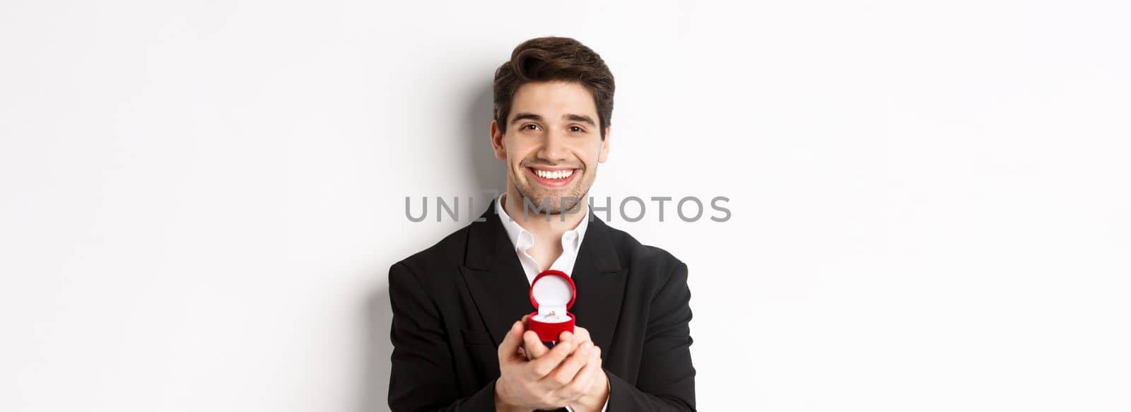 Image of handsome man looking romantic, open small box with engagement ring, making a proposal and smiling, standing against white background.