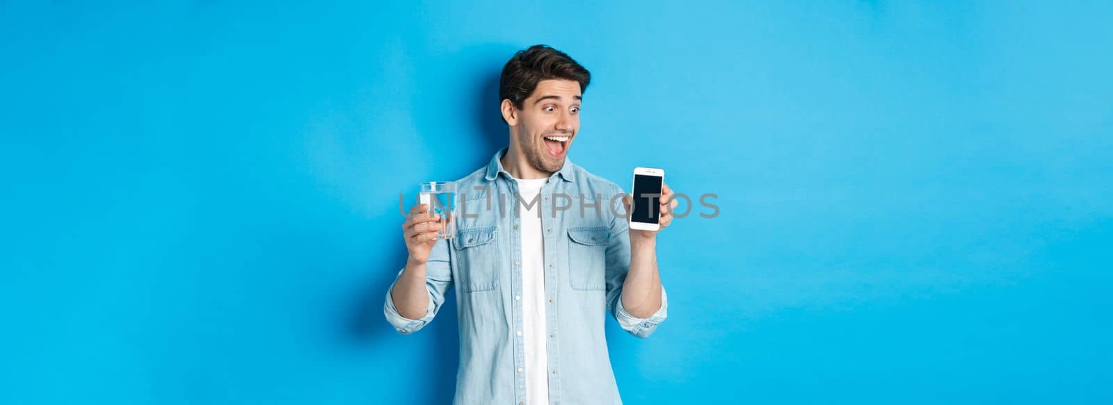 Happy man looking excited at mobile phone screen, holding glass of water, standing over blue background.