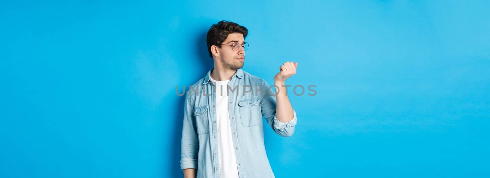 Young handsome man in glasses looking at his fingernails, checking manicure, standing over blue background.