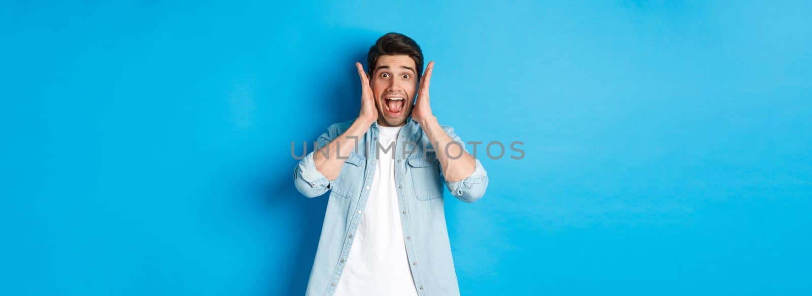 Scared man screaming and looking startled at something, standing against blue background.