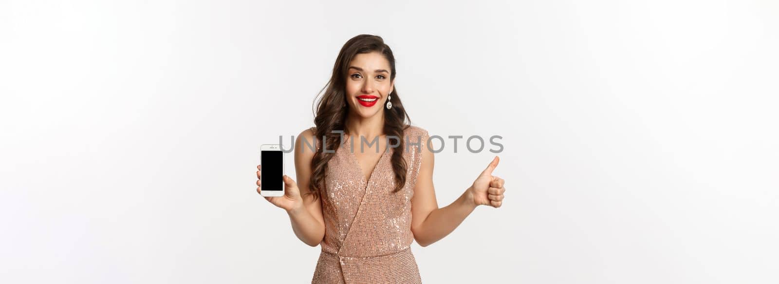 Online shopping. Beautiful young woman in party dress and makeup, showing thumbs up and mobile screen, praising good app, white background.