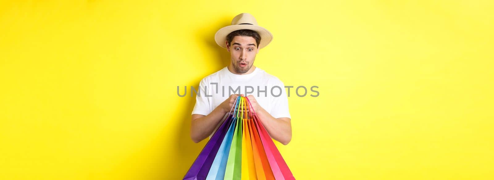Happy man looking surprised at shopping bags, buying souvenirs on vacation, standing over yellow background.
