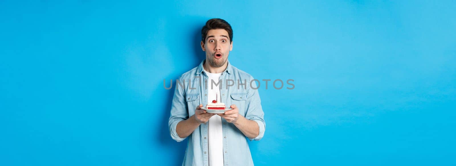 Man holding birthday cake and looking surprised, making a wish on lit candle, standing over blue background.