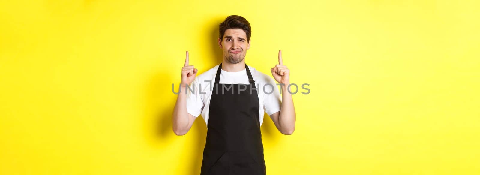Displeased and skeptical waiter in black apron pointing fingers up, express dislike and indifference, standing over yellow background by Benzoix