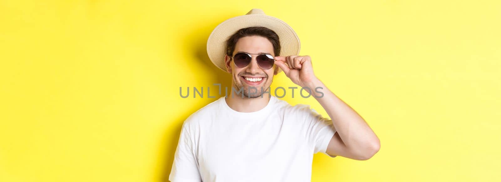 Concept of tourism and vacation. Close-up of handsome man tourist looking happy, wearing sunglasses and summer hat, standing over yellow background.