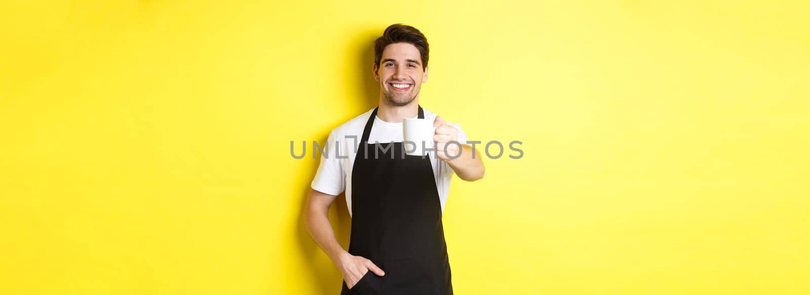 Handsome barista in black apron giving you cup of coffee and smiling, standing over yellow background by Benzoix