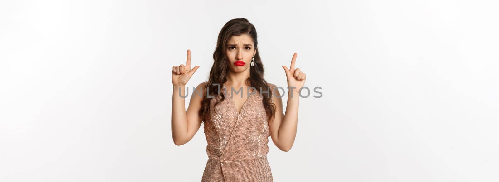 New Year, christmas and celebration concept. Gloomy and sad glamour girl in party dress pointing fingers up, sulking and crying about something, standing over white background by Benzoix