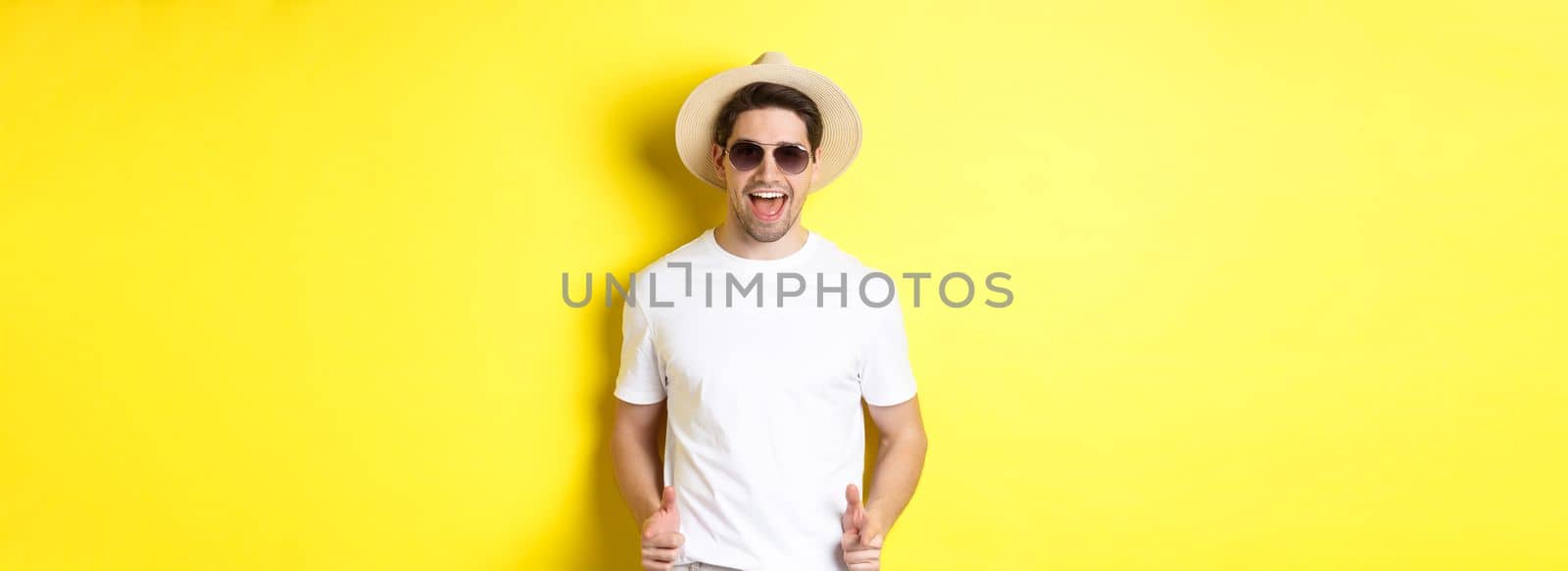 Confident and cheeky guy on vacation flirting with you, pointing finger at camera and winking, wearing summer hat with sunglasses, yellow background.