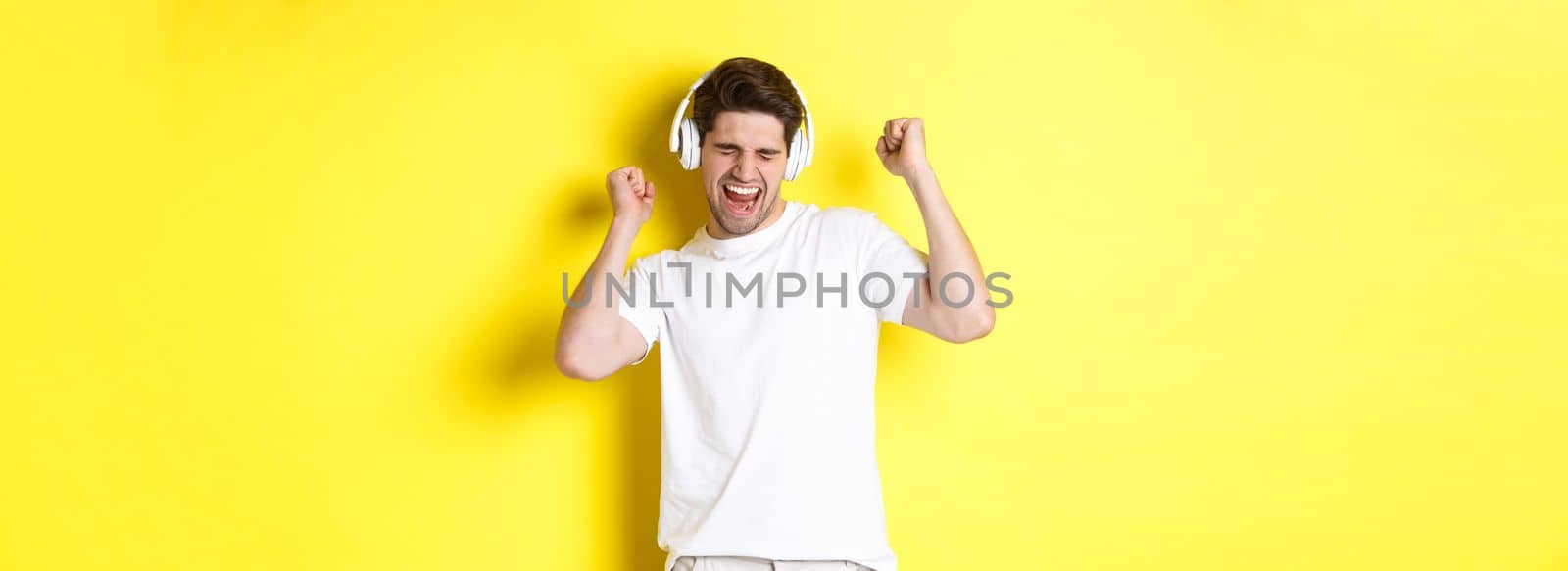 Happy man dancing and listening music in white headphones, standing over yellow background. Copy space