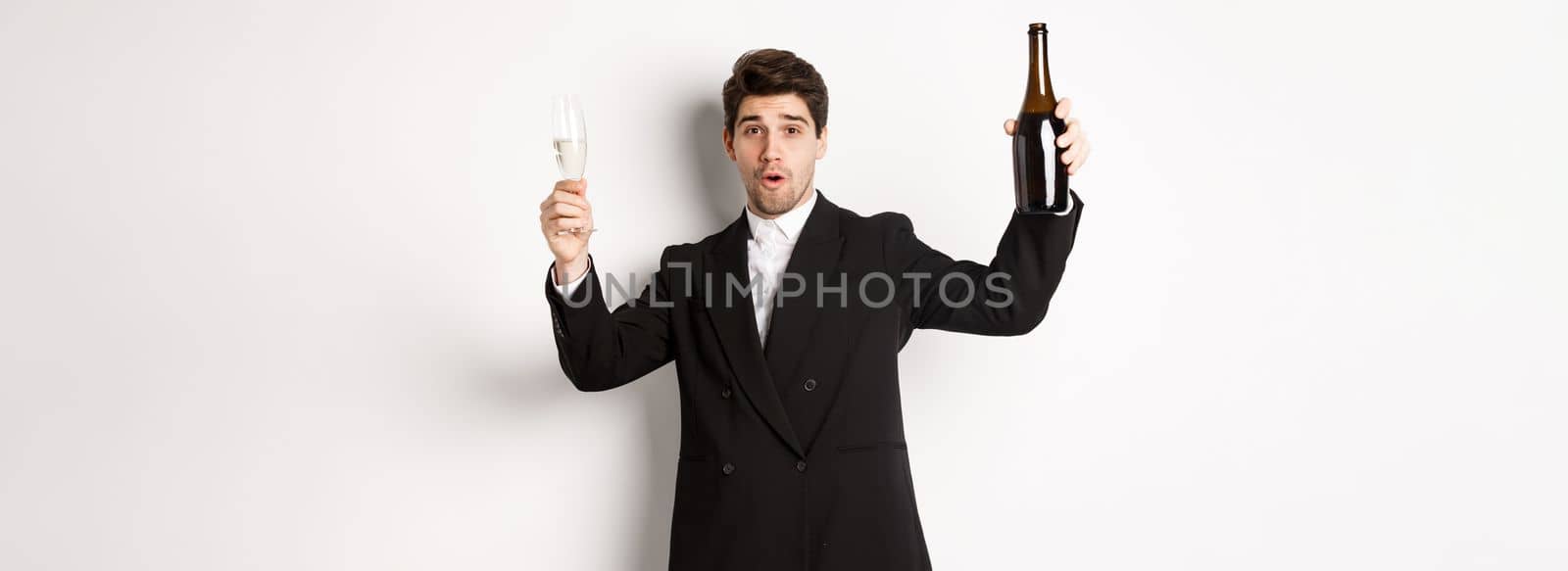 Concept of holidays, party and celebration. Image of handsome man in stylish suit, dancing with bottle of champagne, drinking on new year, standing over white background.