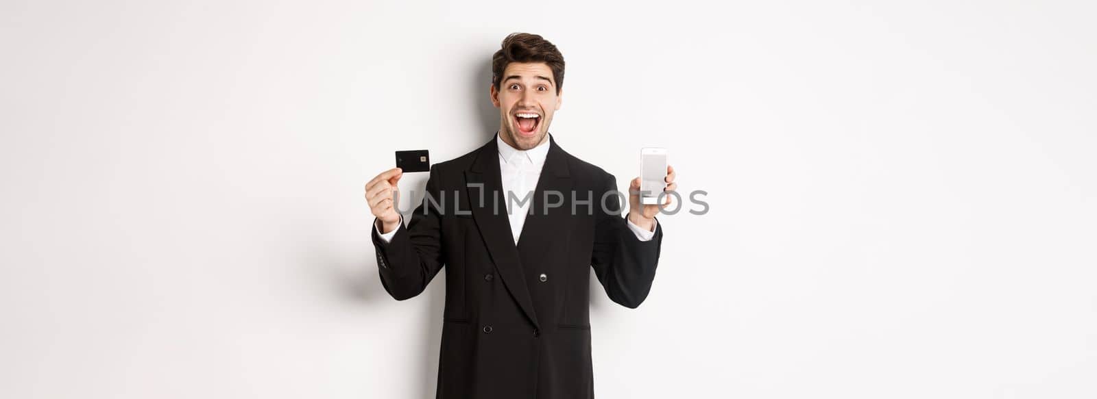 Image of handsome businessman in black suit, looking excited and showing credit card with mobile phone screen, standing against white background by Benzoix