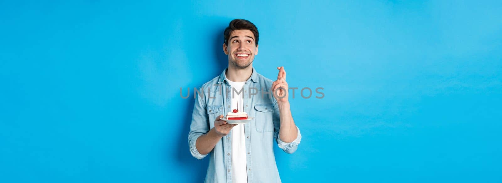 Handsome man making a wish, celebrating birthday, holding b-day cake and cross fingers, standing over blue background.