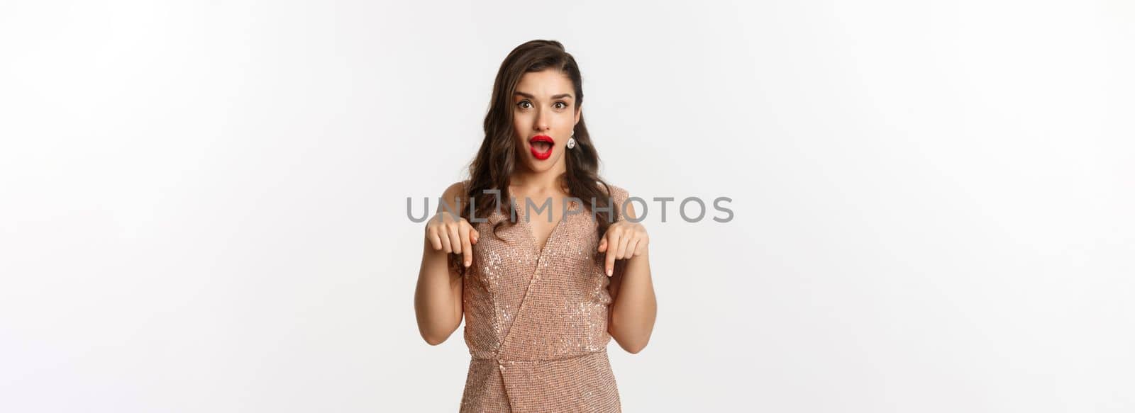 Amazed beautiful woman in glamour dress celebrating christmas party, pointing fingers down at promo, standing over white background.