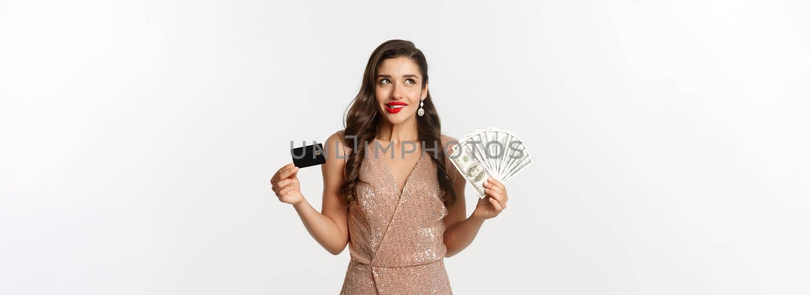 Shopping concept. Young woman deciding what to buy, looking thoughtful at upper left corner and showing credit card with money, standing in party dress.