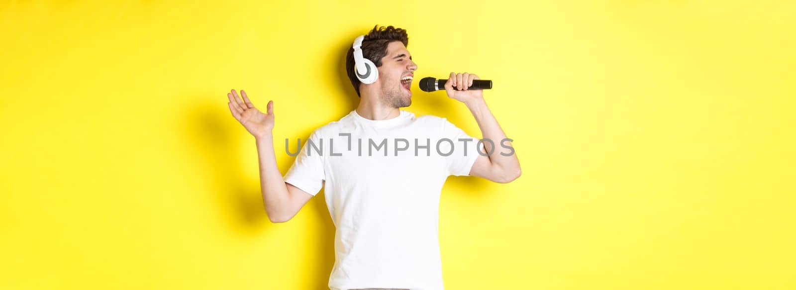 Passionate guy in headphones holding microphone, singing karaoke song, standing over yellow background in white clothes.