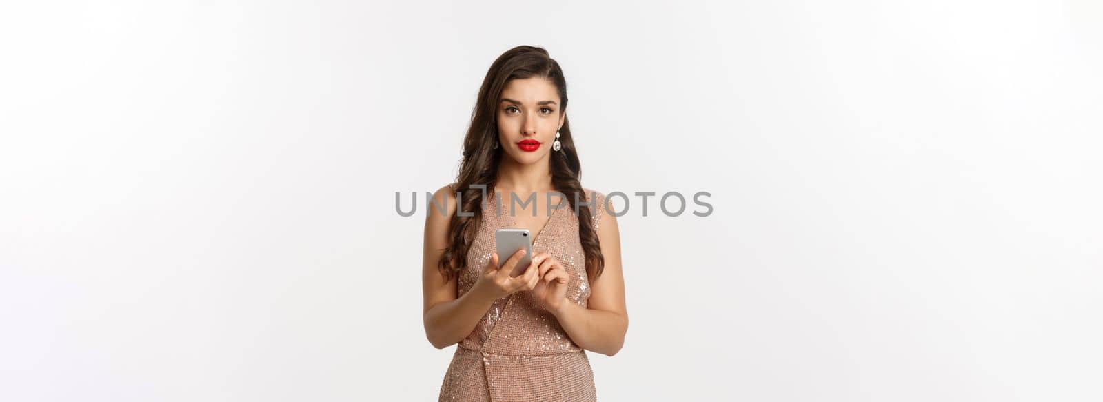 Christmas party and celebration concept. Attractive woman in luxurious dress making phone call, texting message on smartphone, standing over white background.