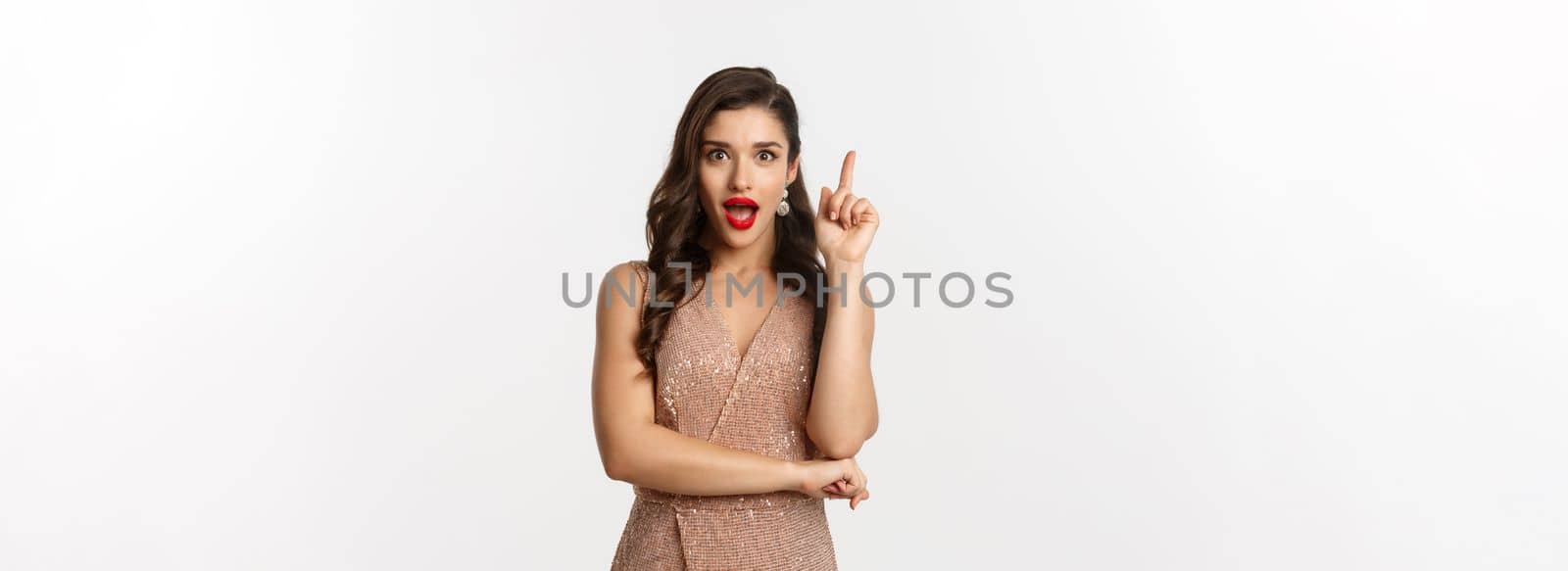 Christmas, holidays and celebration concept. Excited young woman with red lipstick, party dress, having an idea, raising finger in eureka gesture, suggesting something, white background.